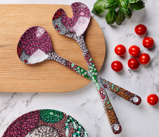 Two melamine salad serving spoons featuring Aboriginal artwork by Coral Hayes Pananka, displayed on a wooden cutting board with cherry tomatoes and basil leaves on a marble surface.