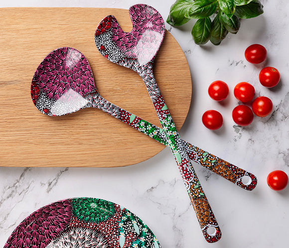 Two melamine salad serving spoons featuring Aboriginal artwork by Coral Hayes Pananka, displayed on a wooden cutting board with cherry tomatoes and basil leaves on a marble surface.