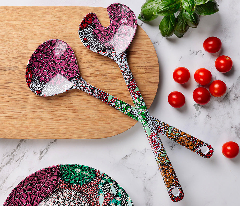 Two melamine salad serving spoons featuring Aboriginal artwork by Coral Hayes Pananka, displayed on a wooden cutting board with cherry tomatoes and basil leaves on a marble surface.