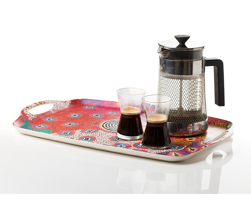 A rectangular serving tray with Aboriginal Art by Ruth Stewart from Warlukurlangu Artists, holding a stainless steel French press and two small clear glasses filled with espresso, on a white reflective surface.