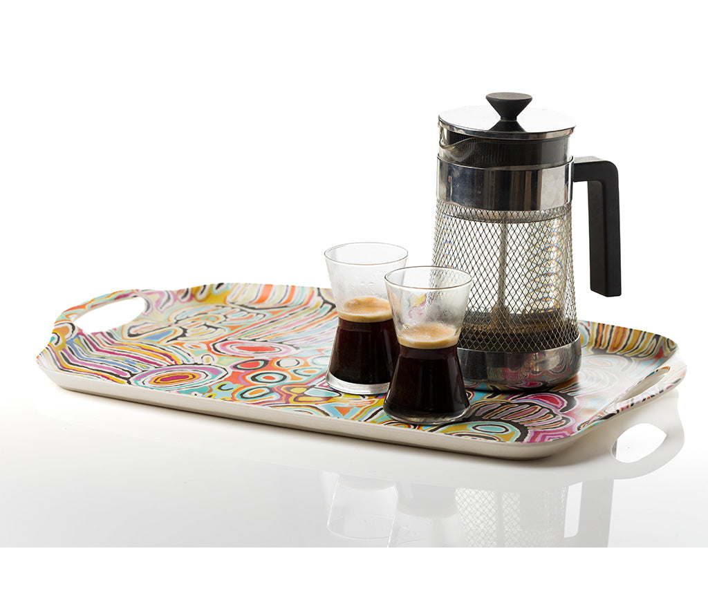 A rectangular serving tray with Aboriginal Art byJudy Napangardi Watson from Warlukurlangu Artists, holding a stainless steel French press and two small clear glasses filled with espresso, on a white reflective surface.