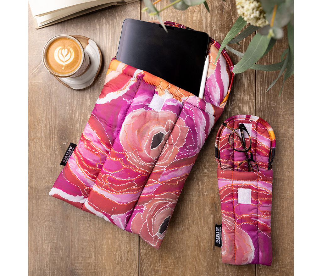 A quilted fabric tablet sleeve with Aboriginal Artwork by Steven Nelson from Warlukurlangu Artists, shown with a tablet inside and a smaller folded view of the sleeve on a wooden tabletop next to a cup of latte and greenery.