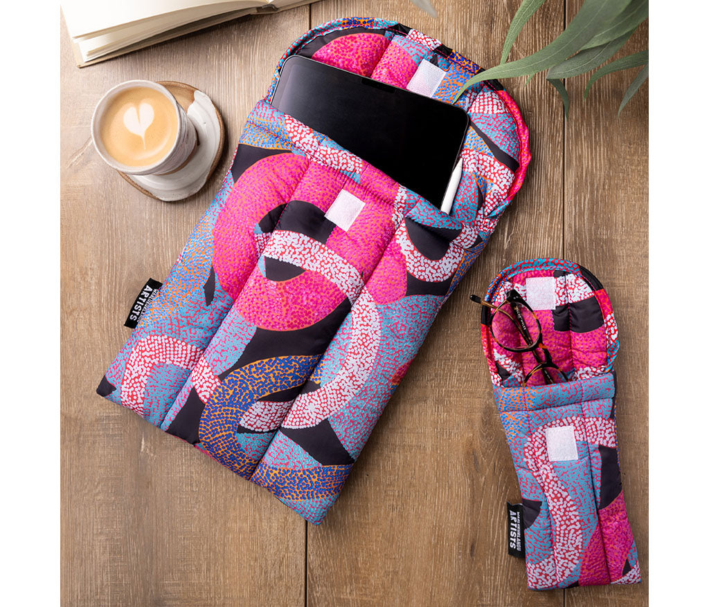 Soft Padded tablet sleeve with a matching small pouch, styled on a wooden tabletop with a cup of coffee, an open book, and a plant leaf. Features Aboriginal artwork by Vanetta Hudson from Warlukurlangu Artists
