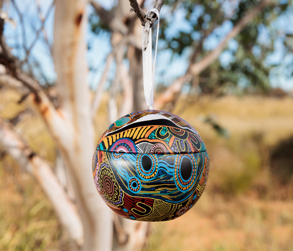 Colorful ornament with Aboriginal design by Justin Butler hanging from a tree branch outdoors.