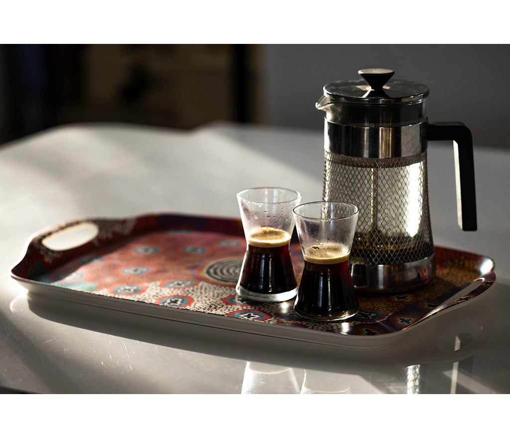 A rectangular serving tray with Aboriginal Art by Ruth Stewart from Warlukurlangu Artists, holding a stainless steel French press and two small clear glasses filled with espresso, on a white reflective surface.